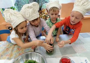 Dzieci ubrane w fartuszki i kucharskie czapki, wkładają do słoika ogórki.
